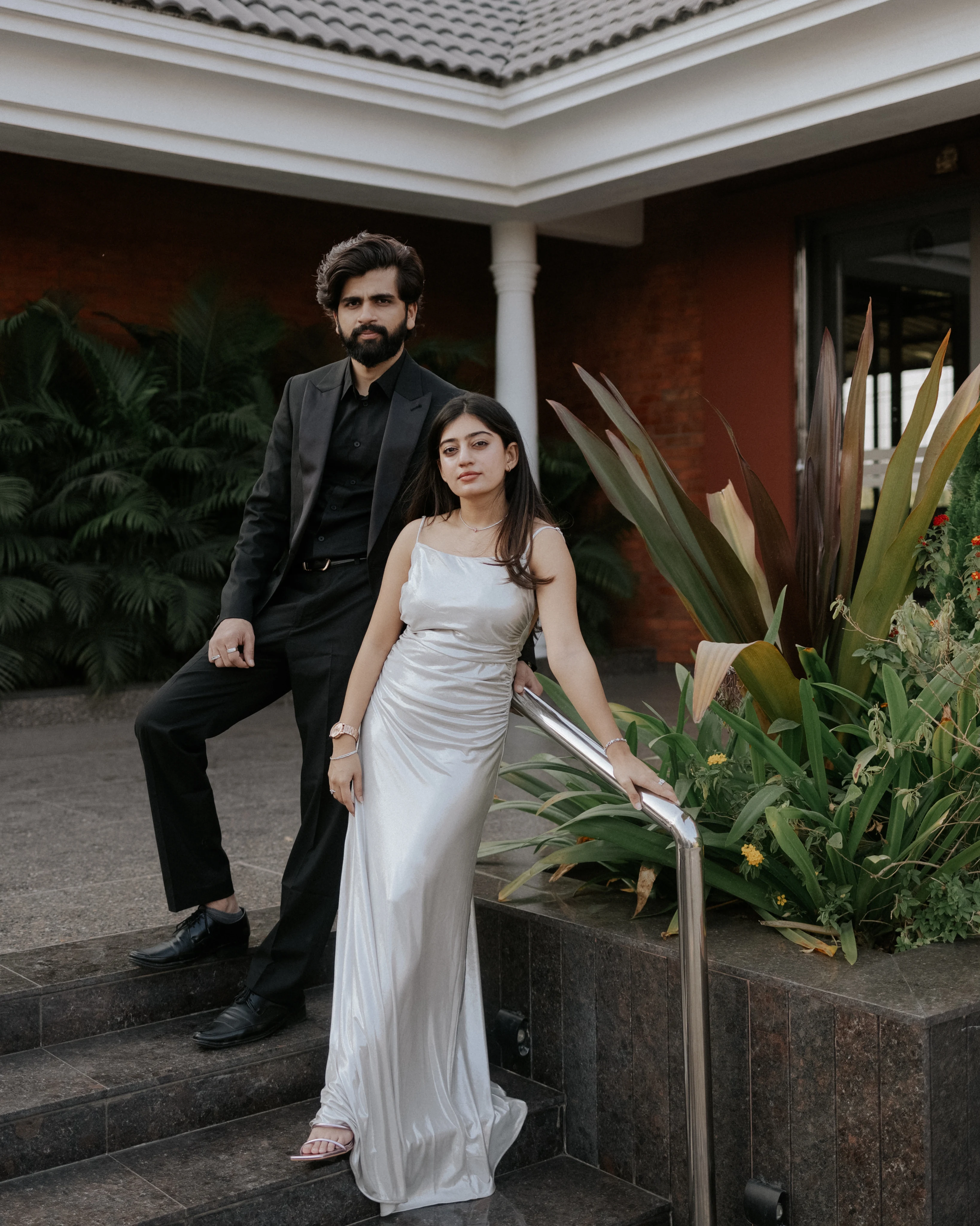 Couple in a black suit and silver satin gown posing on stone steps beside a tropical garden