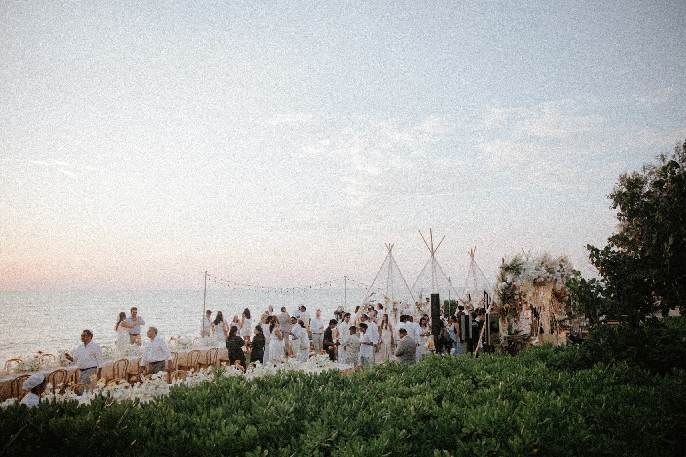Wedding guests gathered at a seaside reception with tipis at dusk